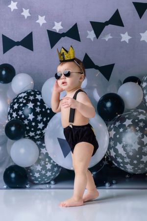 Small child in black outfit and crown strikes pose with balloon decorations and bow tie backdrop.