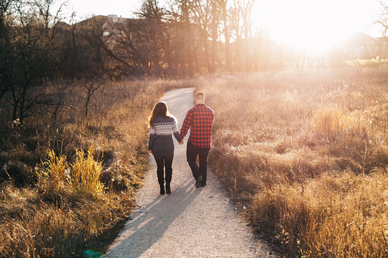 Couple holding hands walking down sunlit autumn path surrounded by golden foliage.