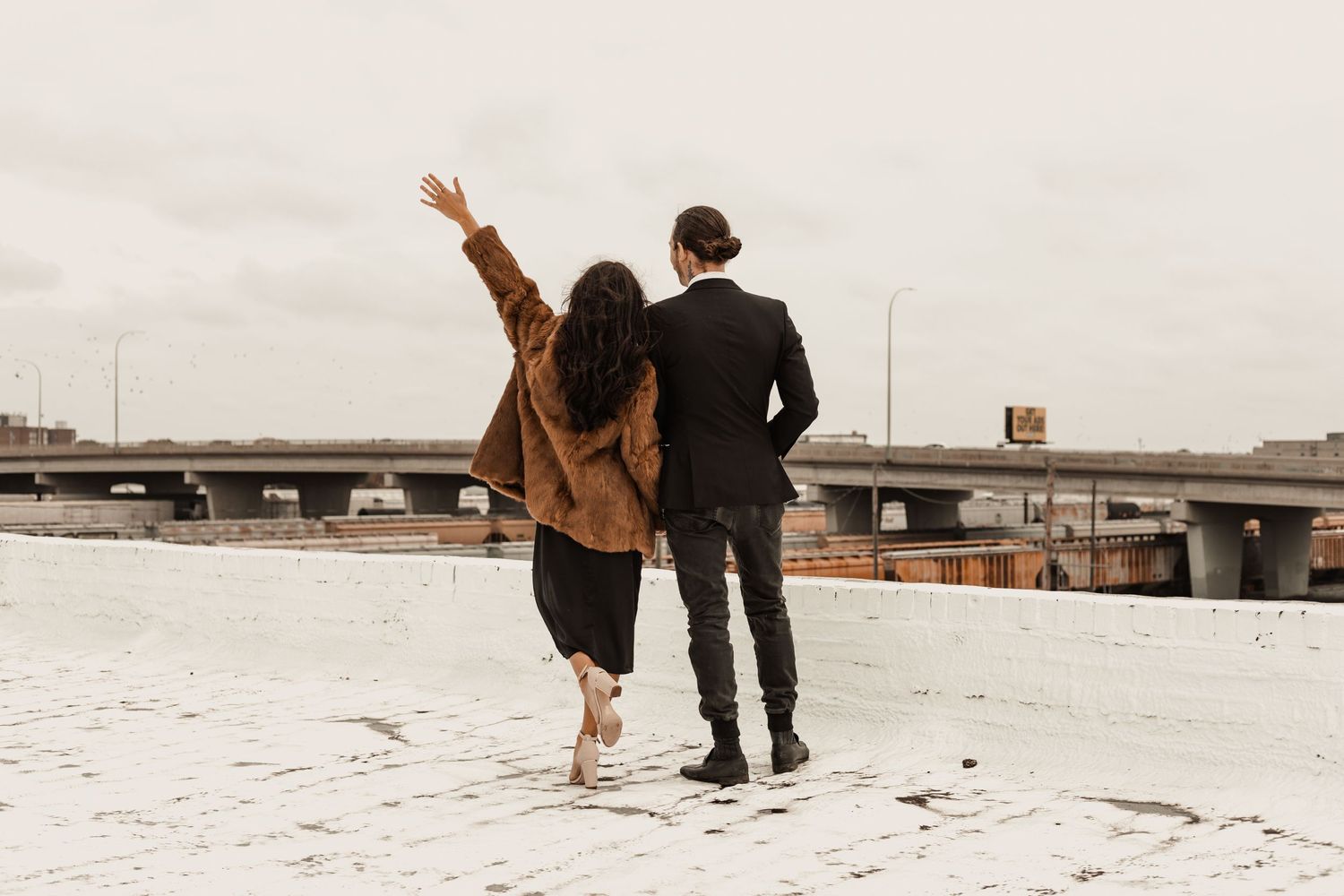 Couple walking together on rooftop, woman raising her arm joyfully, wearing brown jacket and black outfit.