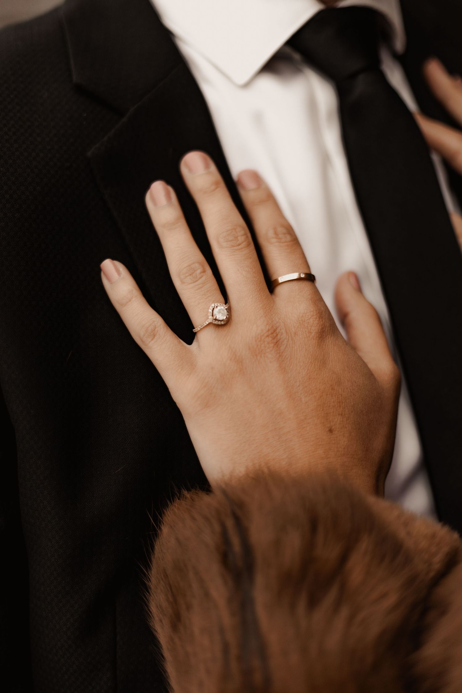 Close-up of a bride's hand with engagement and wedding rings resting on groom's black suit lapel.