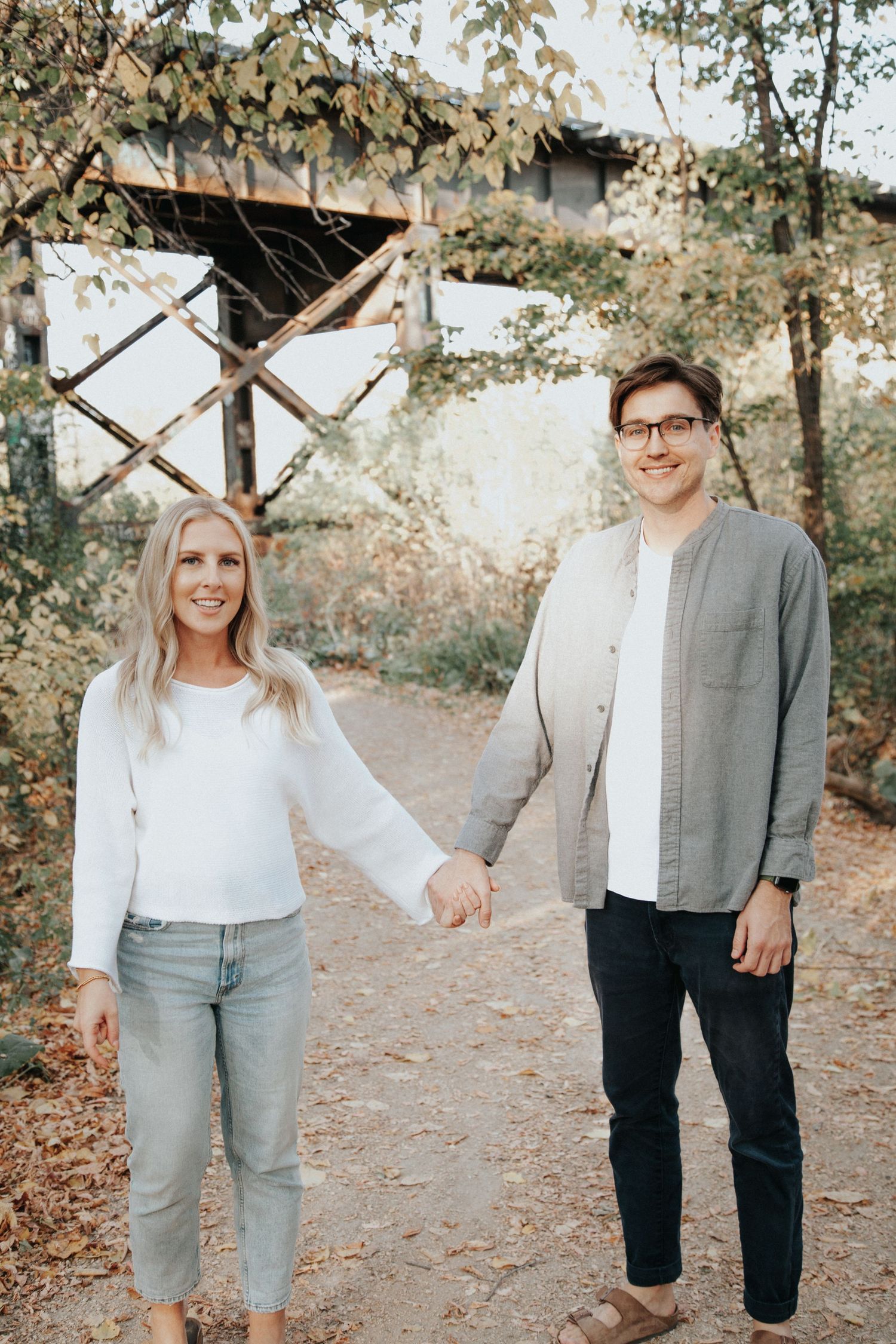 A smiling couple holds hands on a scenic autumn forest path.