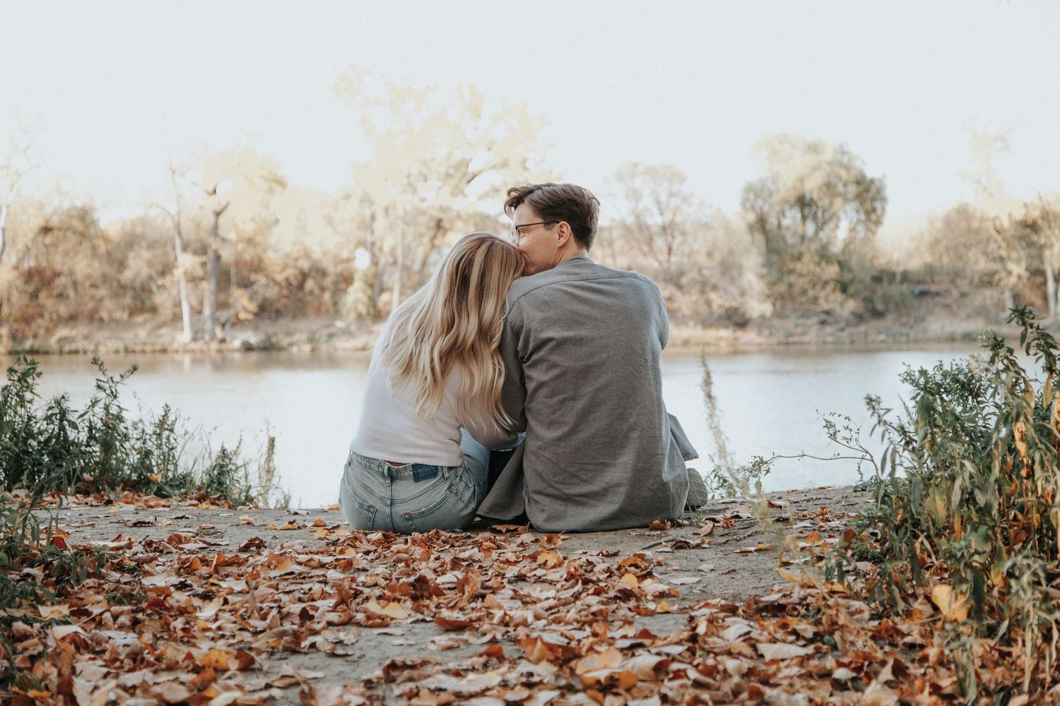 A romantic couple sits together by a peaceful lake surrounded by fall foliage.