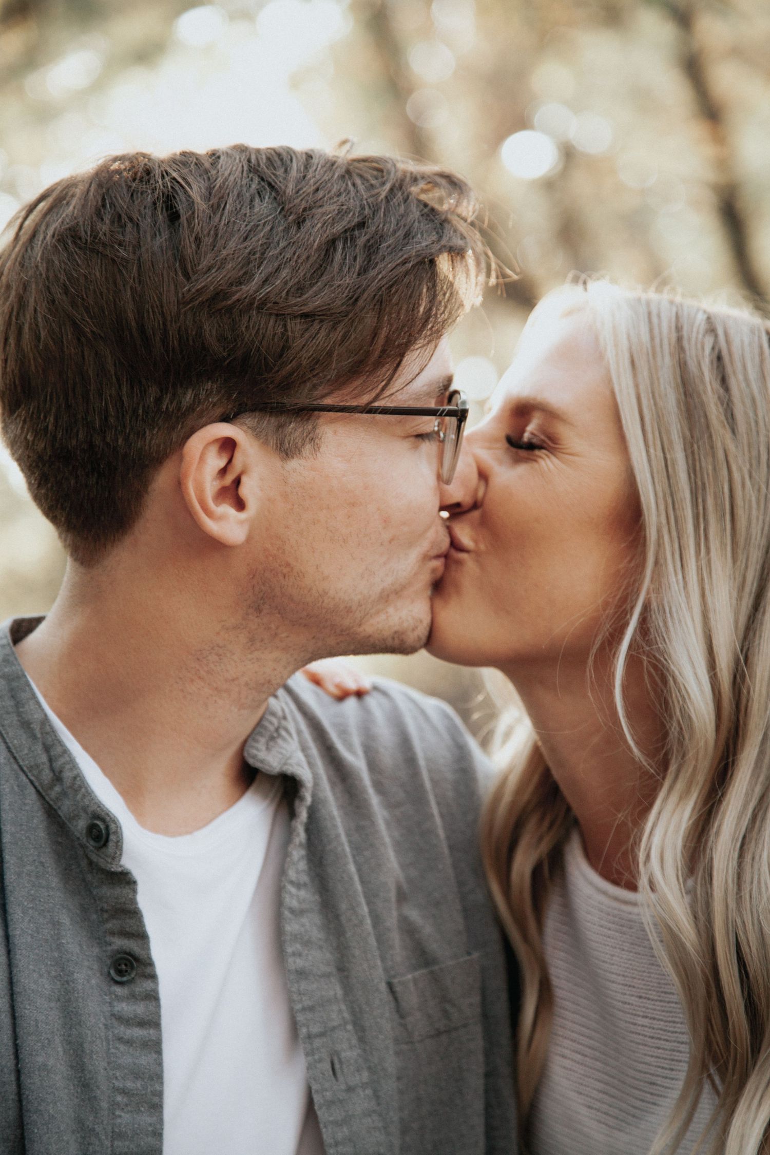 A couple shares a passionate kiss outdoors in a sun-dappled forest setting.