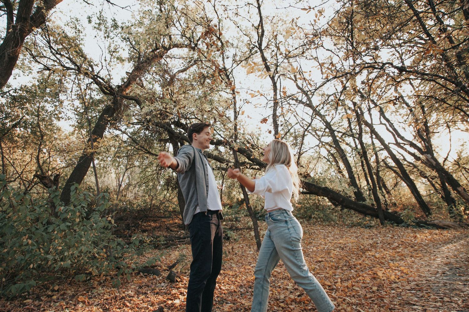 A playful couple dances together on a leaf-covered path in an autumn forest.