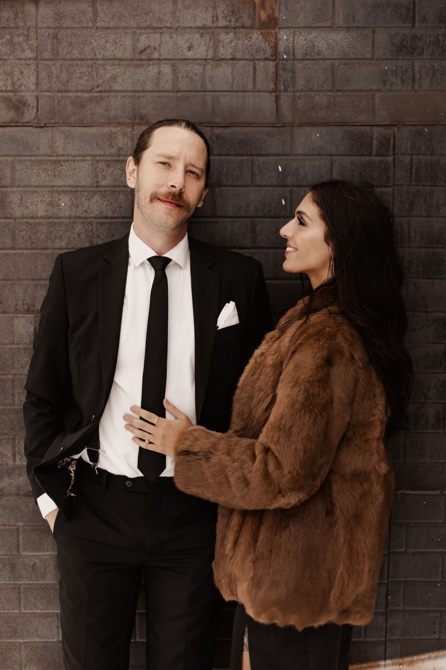 Stylish couple posing against dark brick wall, man in black suit, woman in fur coat.