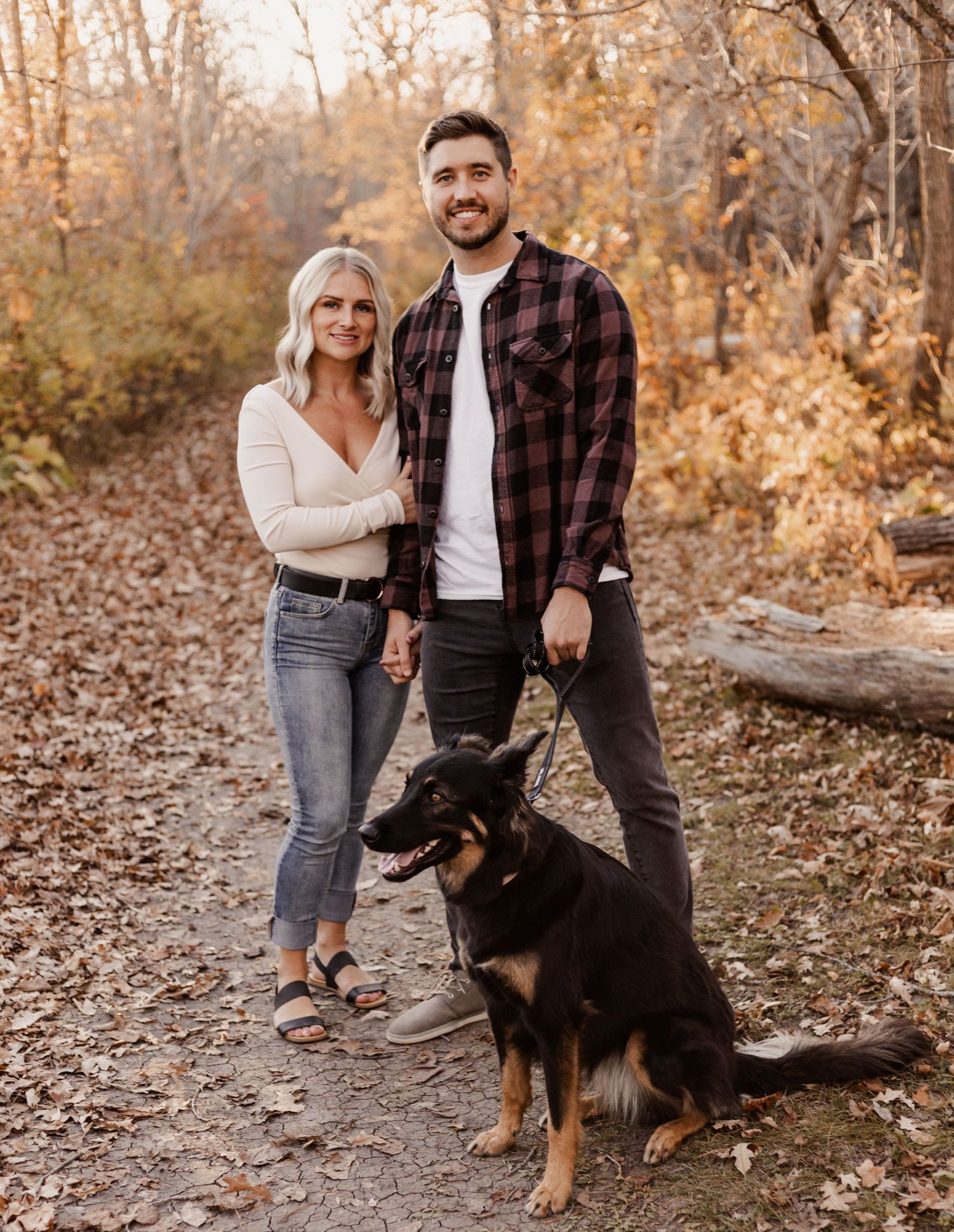 Smiling couple with German Shepherd dog posing on autumn leaf-covered forest path.