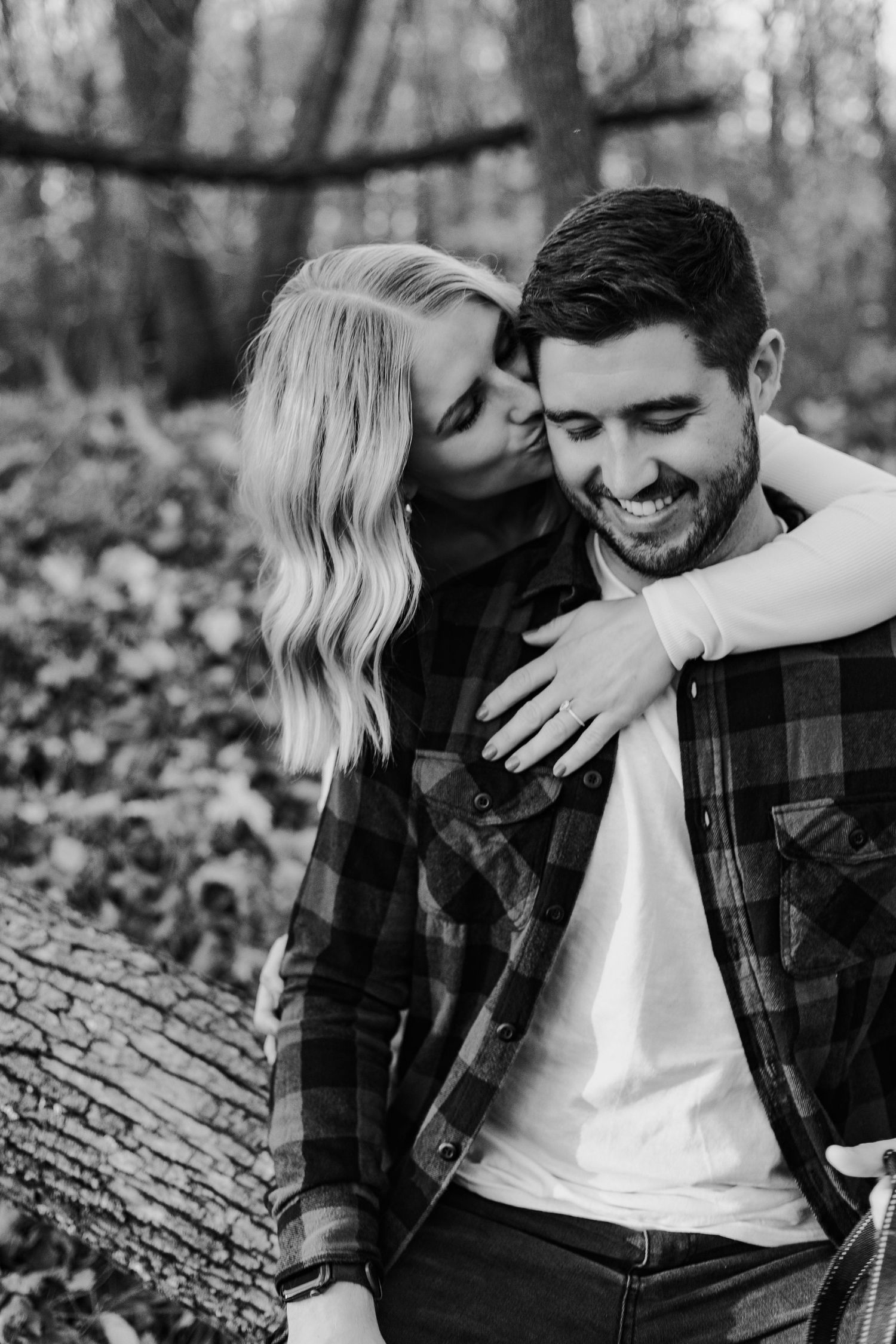 Black and white photo of woman kissing mans cheek while embracing from behind near log.