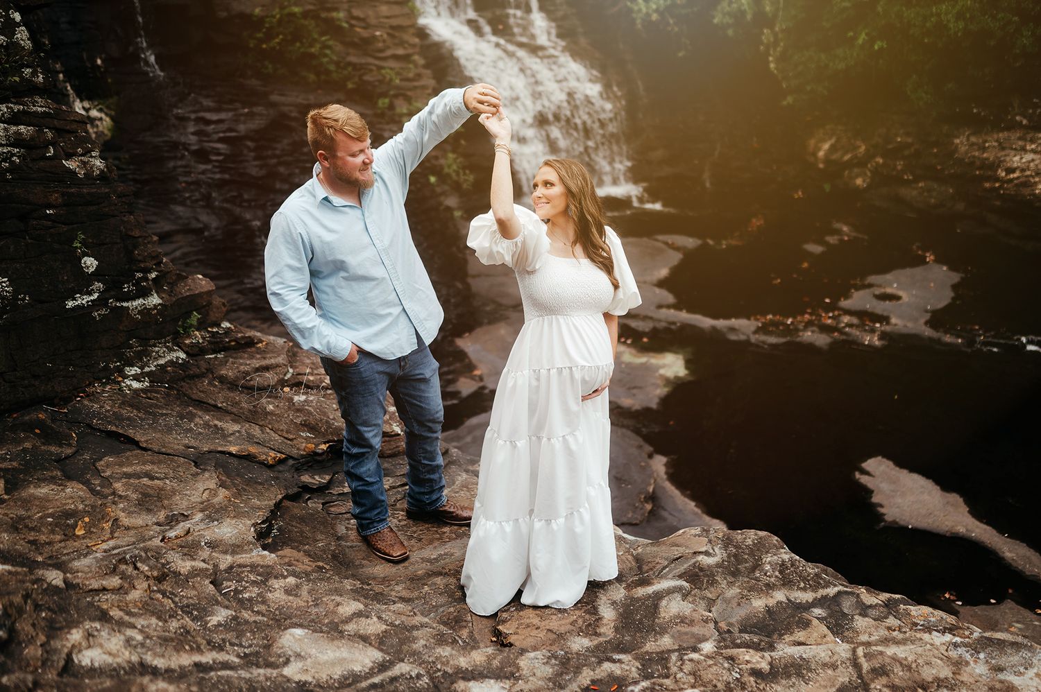 Couple dancing on rocky outcrop near waterfall during engagement session.