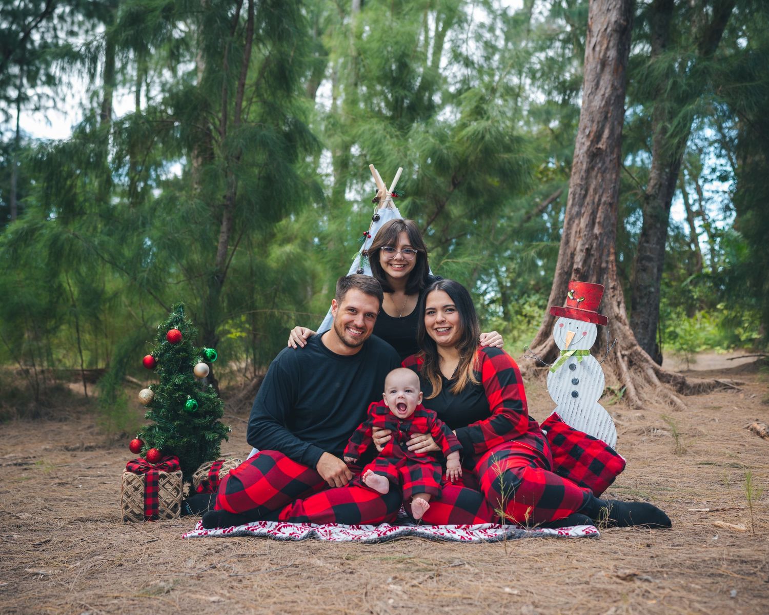 Family wearing matching buffalo plaid pajamas posed by Christmas tree and decorations in forest setting.