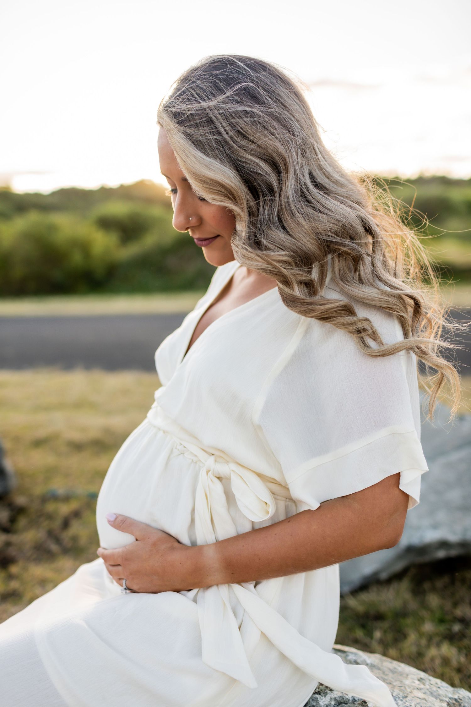 A woman in a flowing white dress cradles her baby bump against a sunset backdrop.