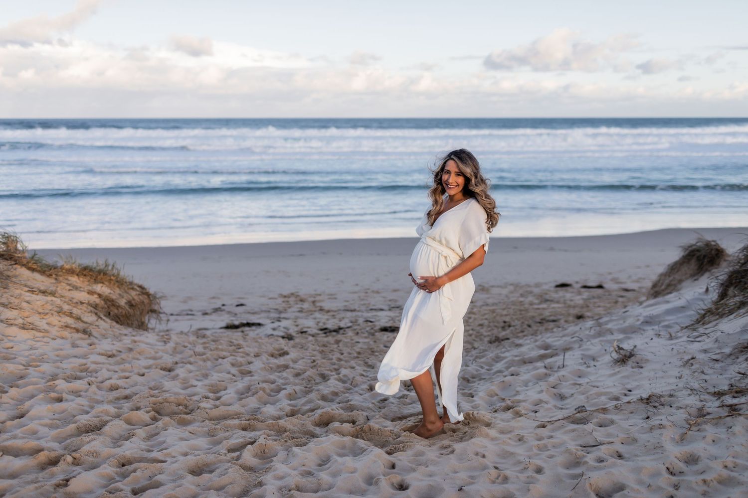 A woman in white dress stands on a beach, holding her pregnant belly during golden hour.