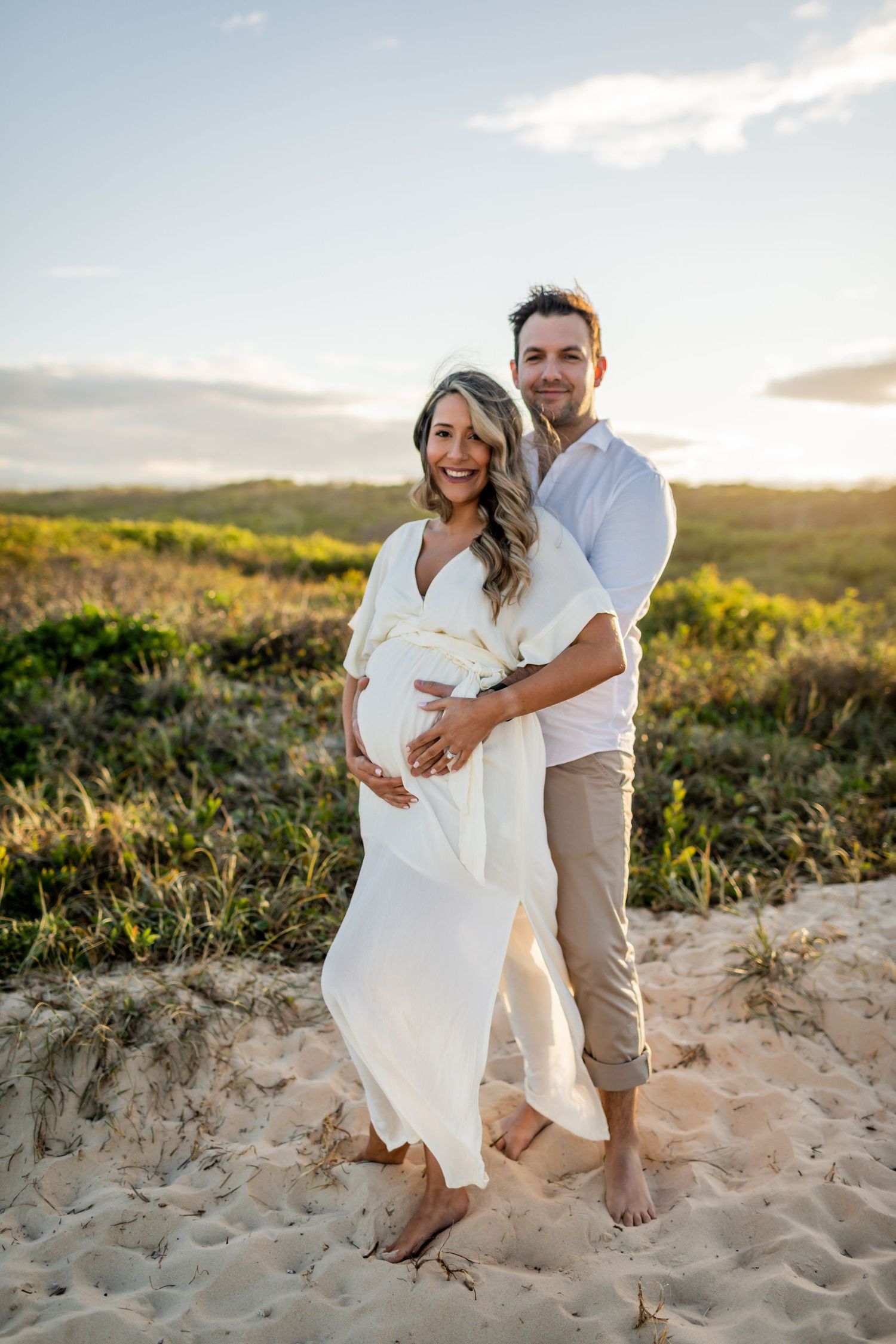A couple embraces on sandy dunes during a sunset maternity photoshoot.