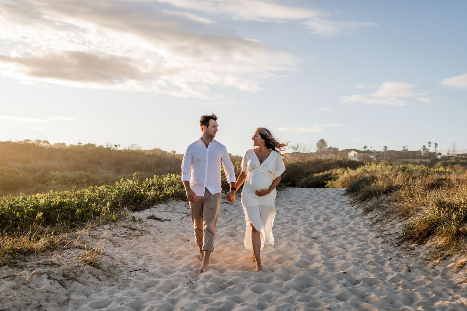 A couple walks hand in hand along a sandy coastal path at sunset.