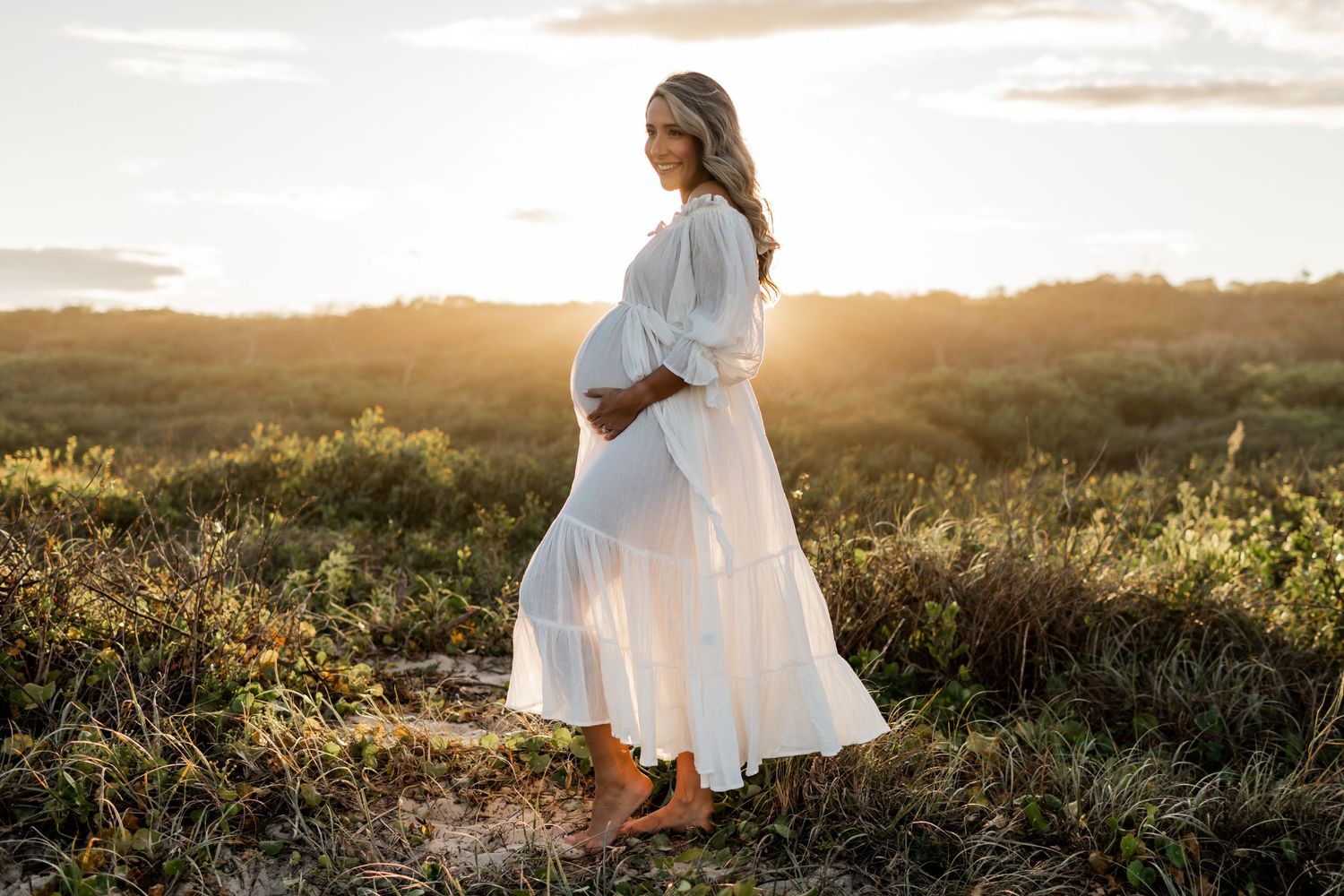 A woman in white dress poses on a hill during golden hour maternity photography.