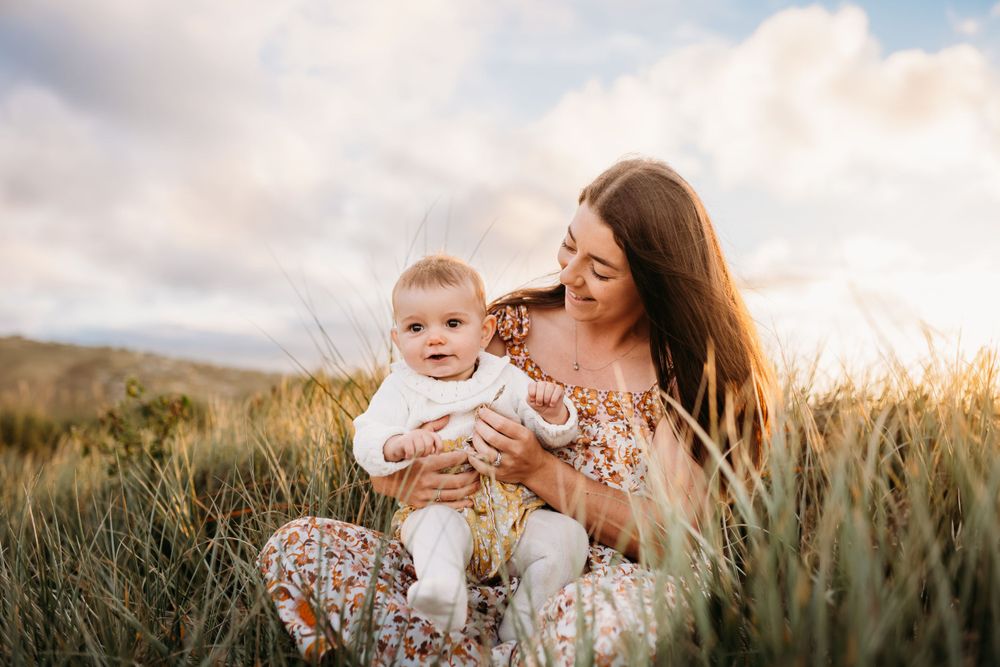 Mother and baby share joyful moments together while sitting in tall grass during golden hour.