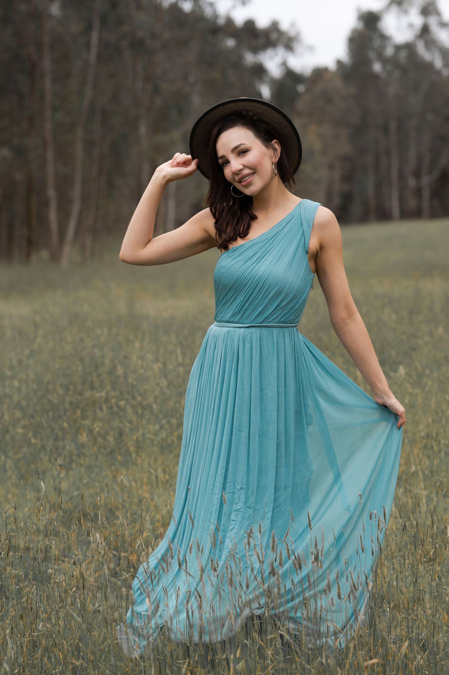 Elegant woman in teal one-shoulder pleated maxi dress and black hat posing in a scenic grassy meadow.