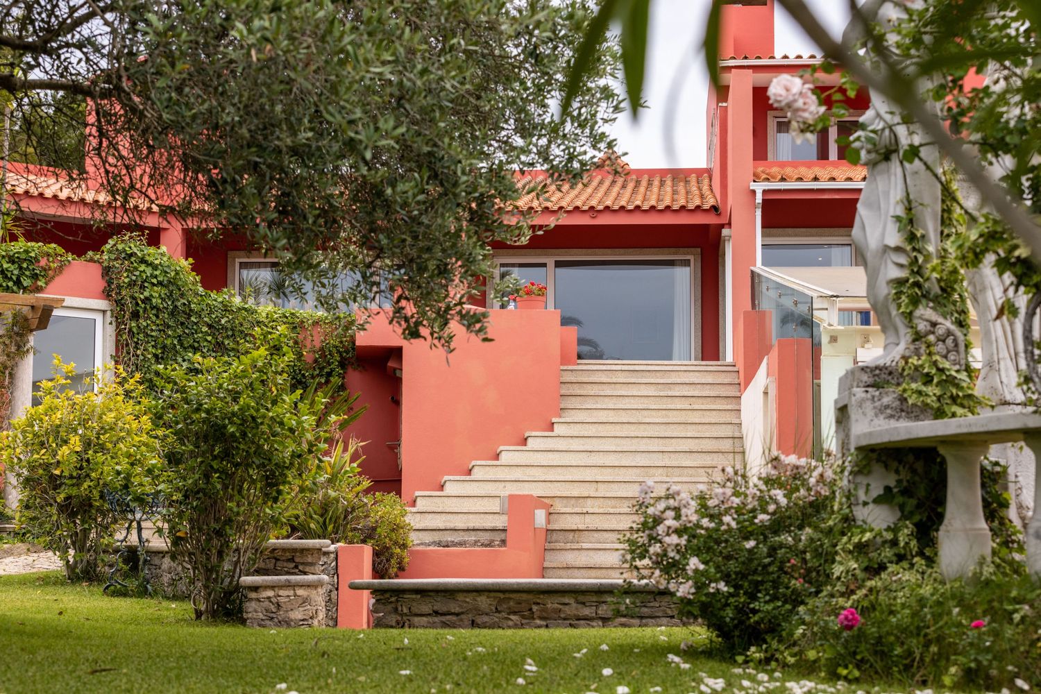 Mediterranean-style pink stucco house with terracotta roof and grand staircase leading to entrance surrounded by lush gardens.