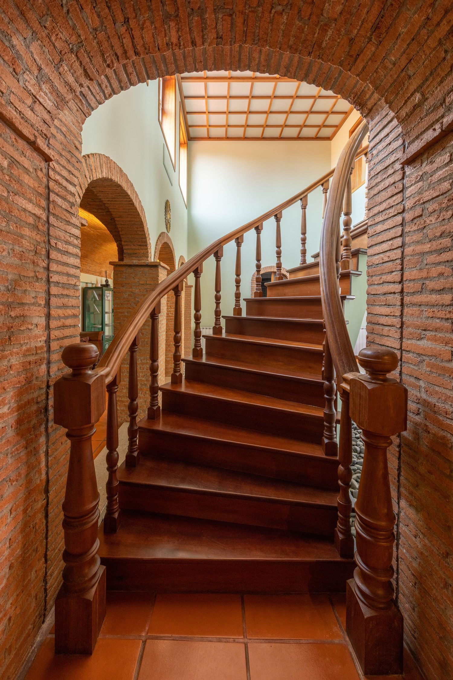 Curved wooden staircase with brick archway and terracotta tiles in Mediterranean style interior.