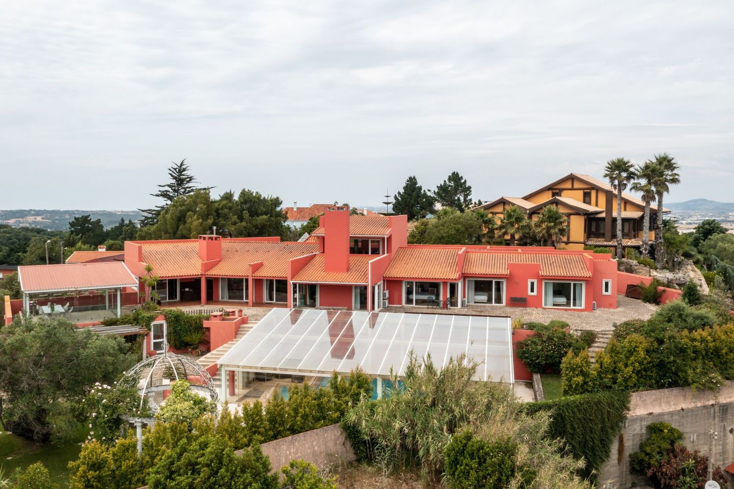 Aerial view of a large pink Mediterranean villa with multiple buildings and terraces nestled in hillside landscape.