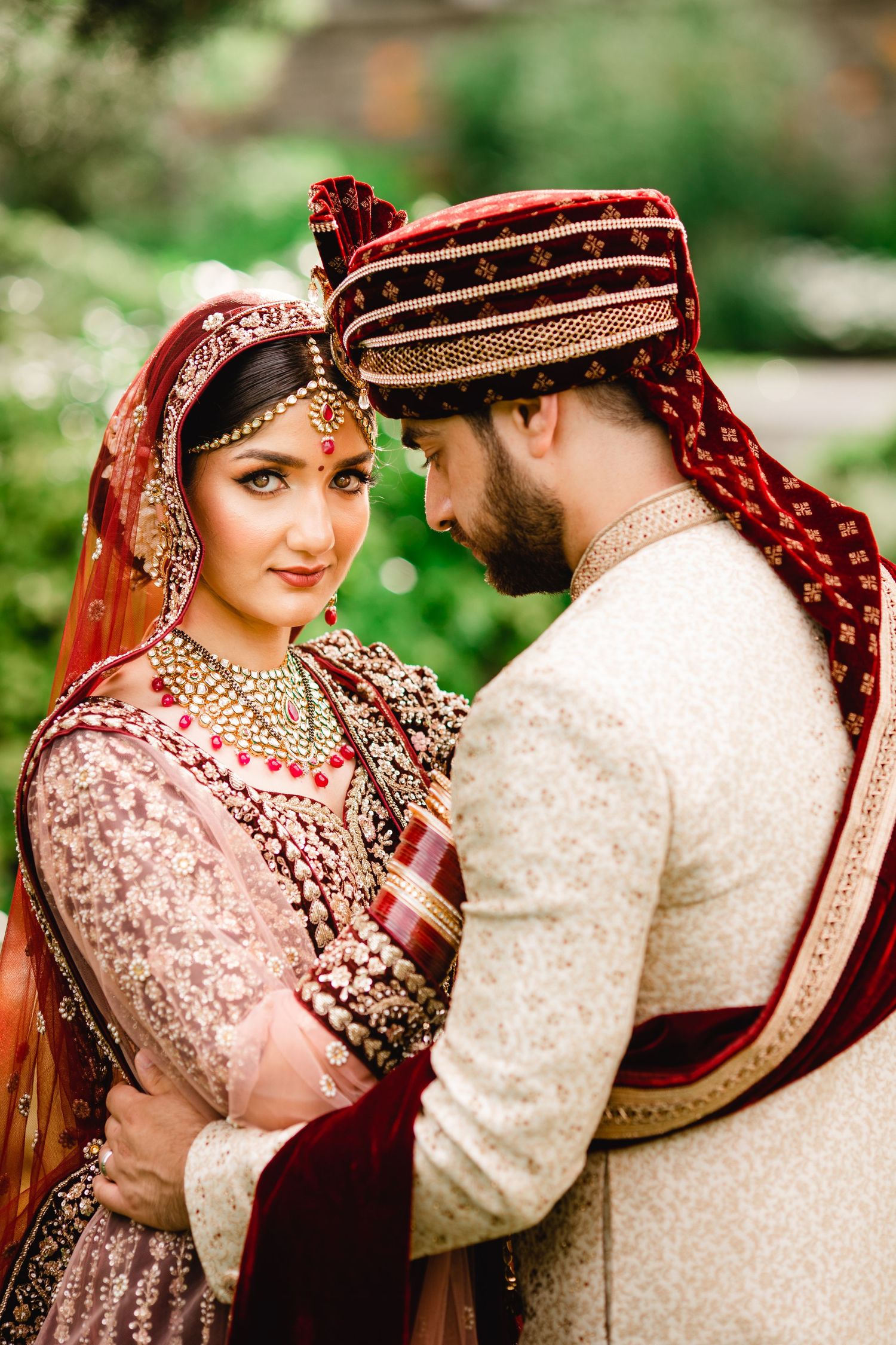 Traditional South Asian wedding couple in ornate red and cream attire share an intimate moment outdoors.