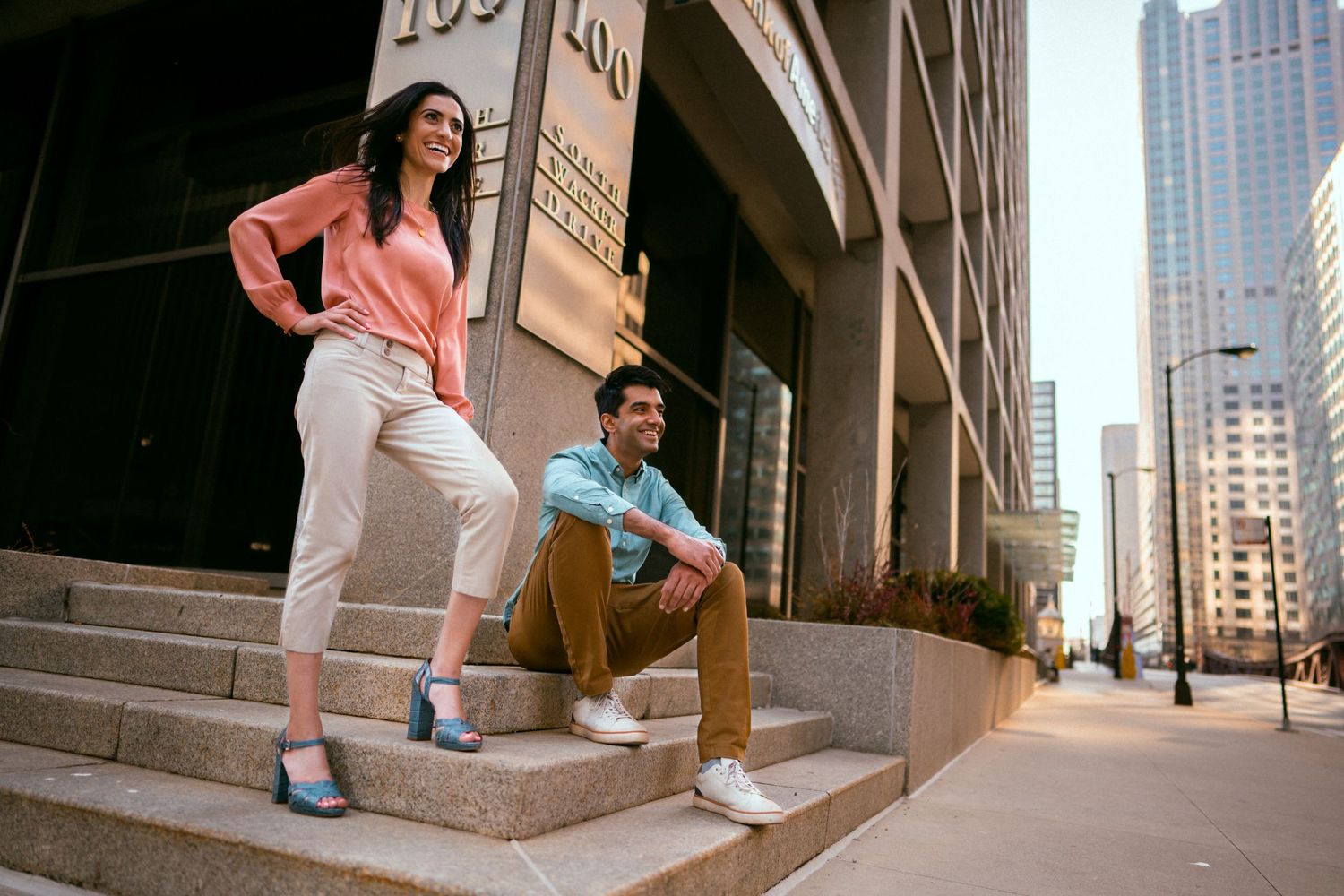Woman stands gracefully, man seated on steps, exuding fashion model allure on a vibrant Chicago street, poised, captivat
