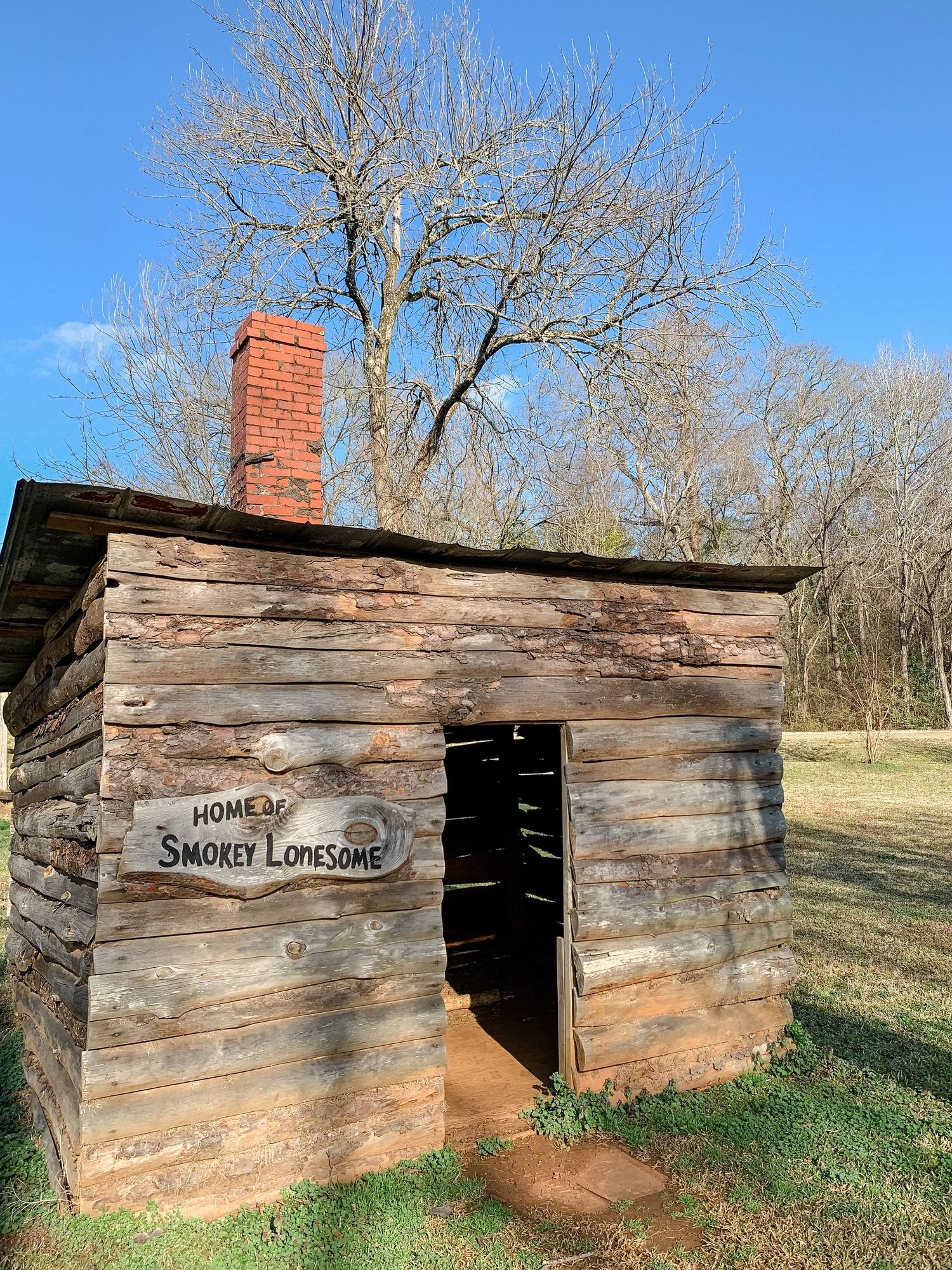 Rustic log cabin structure with red brick chimney in a rural wooded setting.