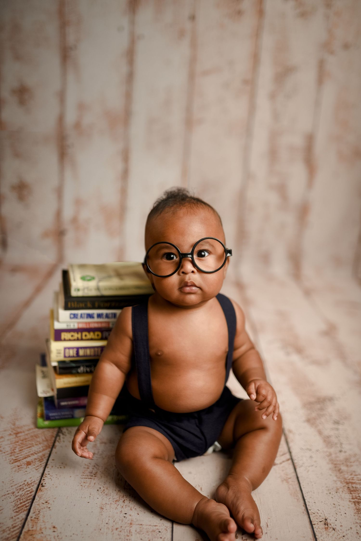 Baby wearing round glasses and suspenders sits against wooden backdrop with stacked books.