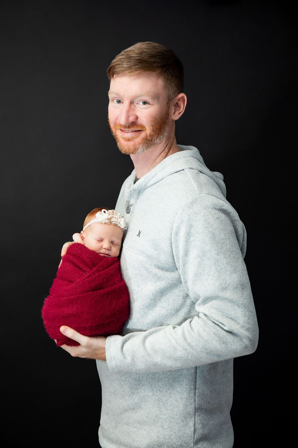 Parent in grey hoodie holds newborn baby wrapped in burgundy against dark background.