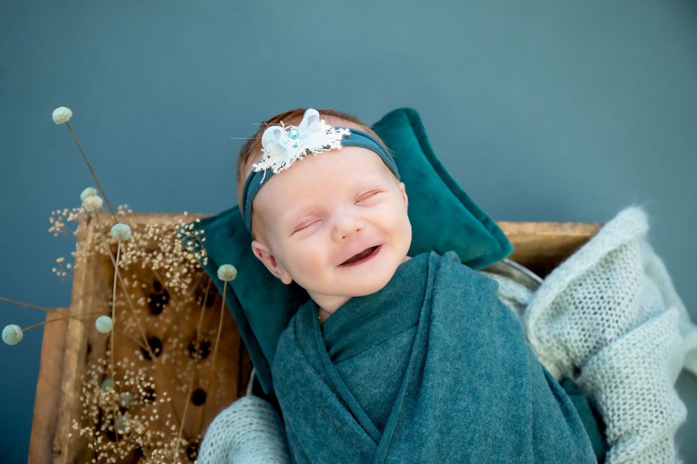 Smiling newborn wrapped in teal fabric lying in wooden box with dried floral accents.