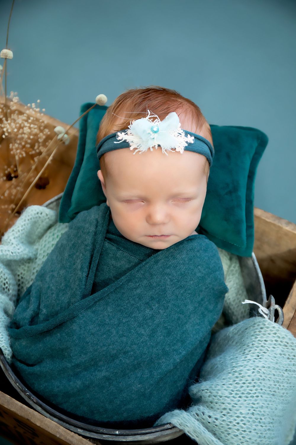 Newborn in teal wrap and floral headband sleeping peacefully against blue backdrop with dried flowers.