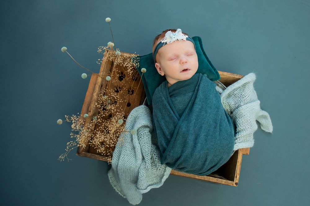 Overhead view of sleeping baby wrapped in teal fabric in wooden box with dried flowers and knit textures.