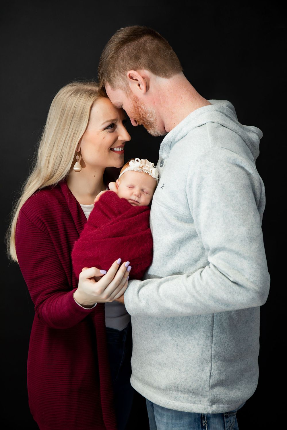 Intimate family portrait with parents holding their newborn wrapped in red against dark backdrop.