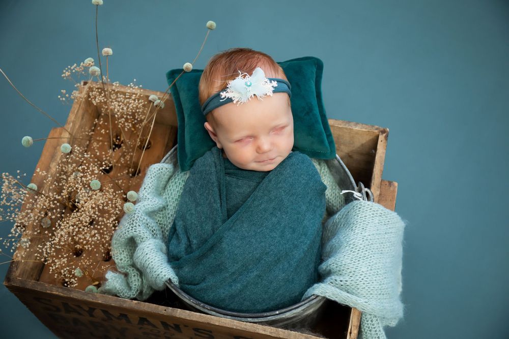 A sleeping newborn swaddled in teal fabric rests in a wooden box with dried flowers and knit accessories.