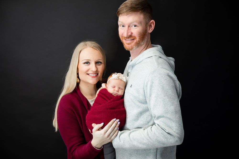 A couple in burgundy and grey holding their newborn baby against a black background for a studio portrait.