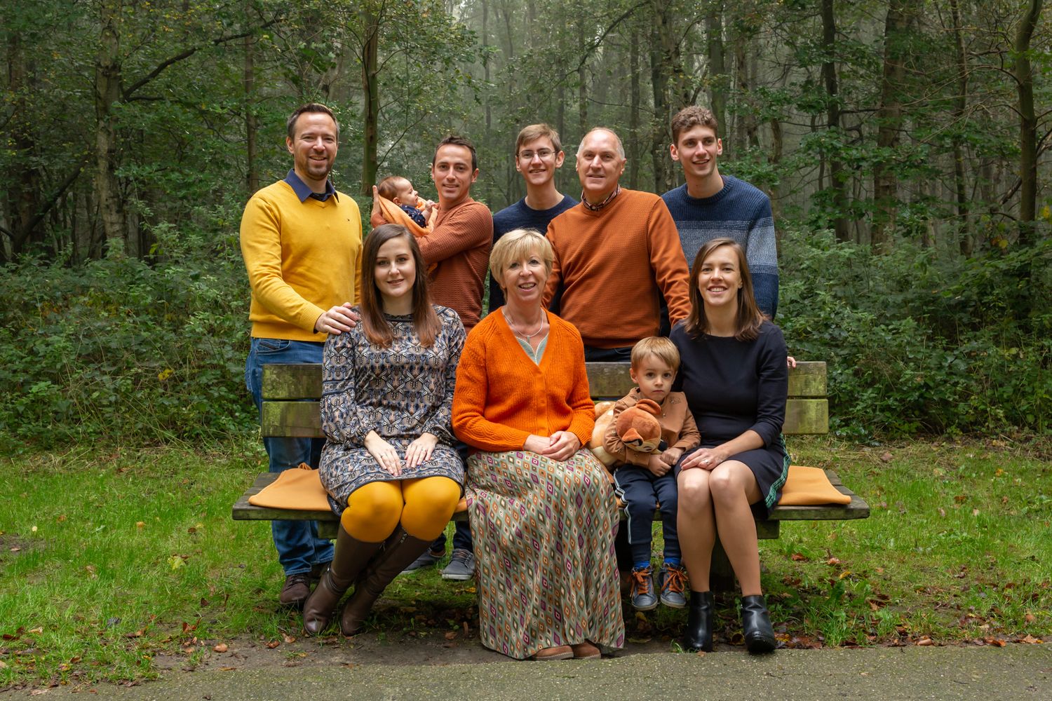 Multi-generational family portrait on wooden bench in lush forest setting.