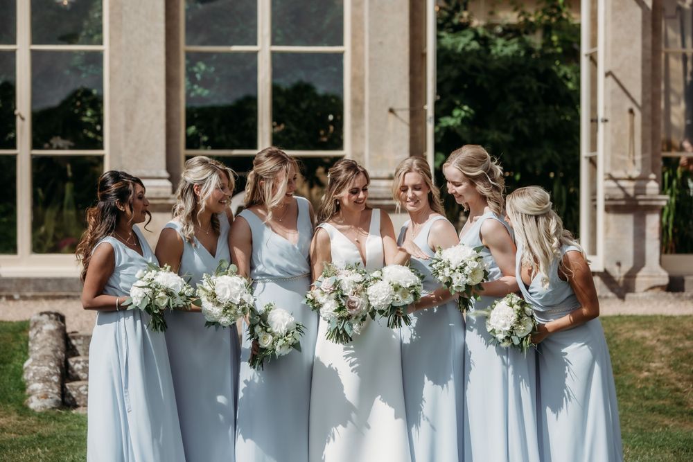 Bridesmaids in light blue dresses holding white bouquets pose by historic building.