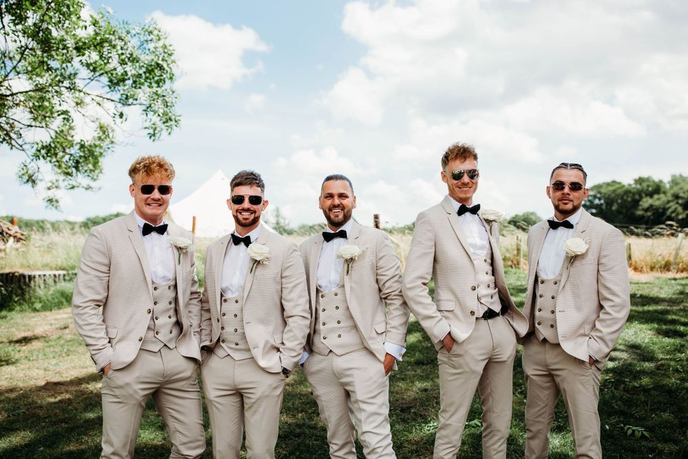 Groomsmen in light beige suits pose together outdoors on sunny wedding day.