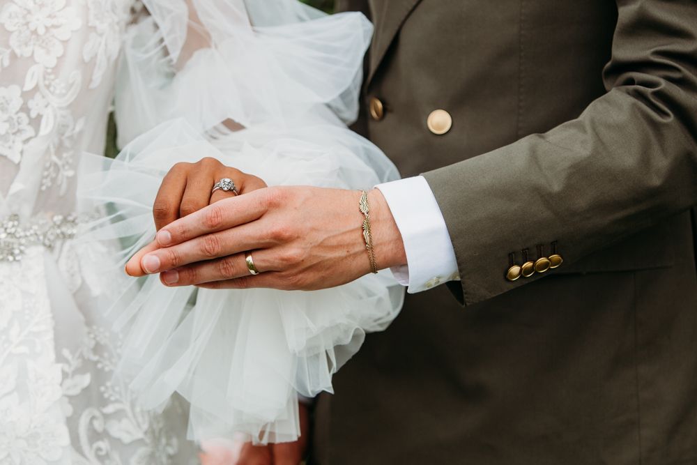 Close-up detail of hands wearing wedding rings against white dress and dark suit fabric.