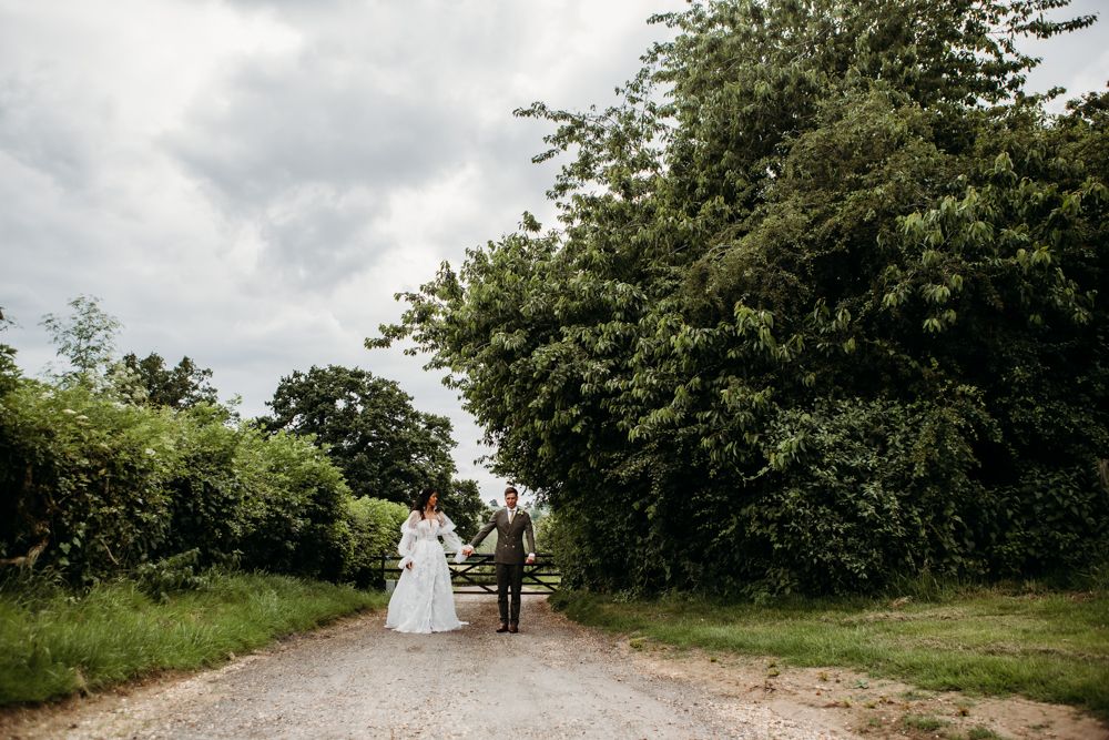 A couple in wedding attire walks down a tree-lined path in the countryside.
