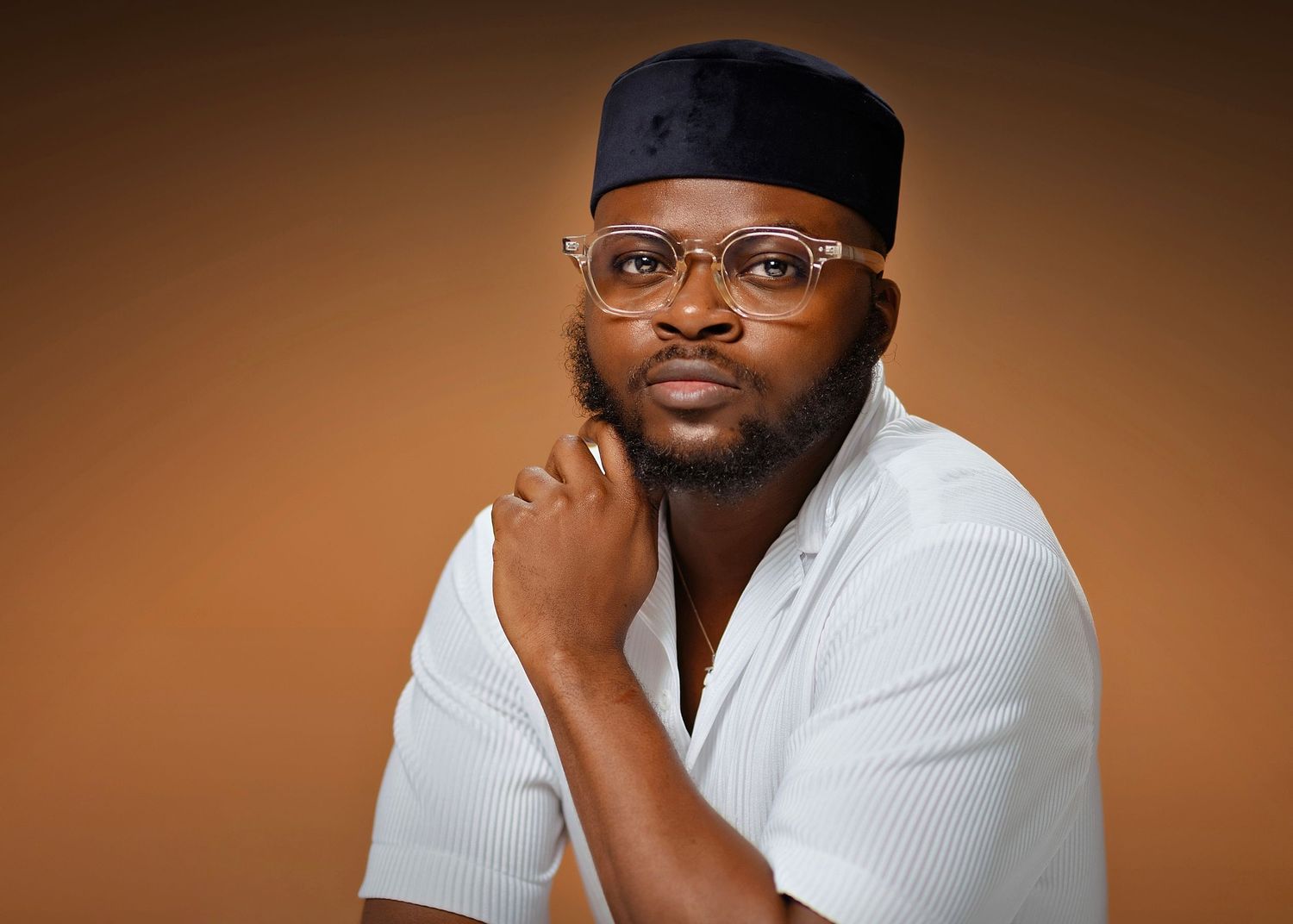 Professional studio portrait against brown background featuring eyeglasses and white shirt with black cap.