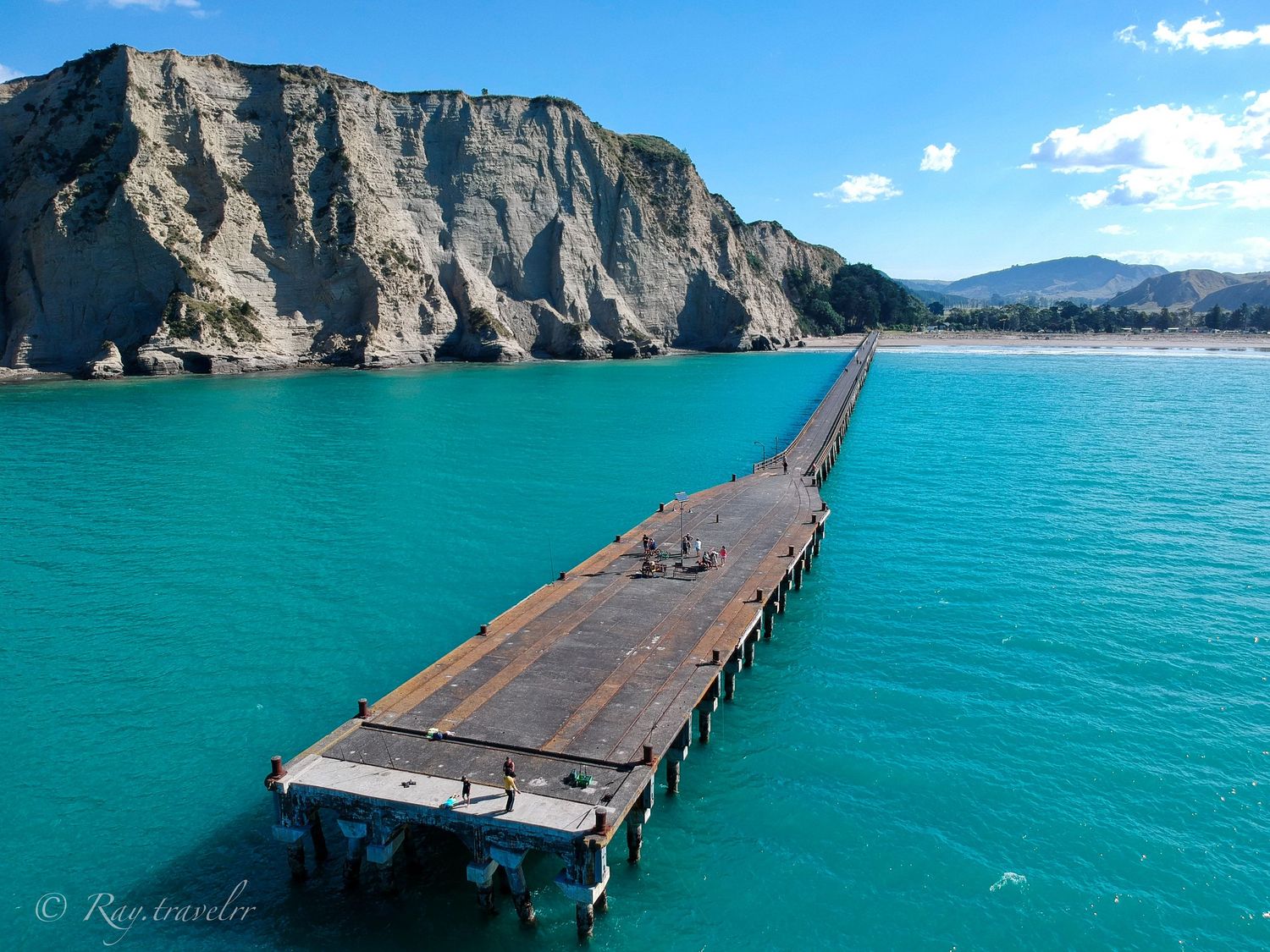 A long wooden pier extends into turquoise waters beneath dramatic coastal cliffs.