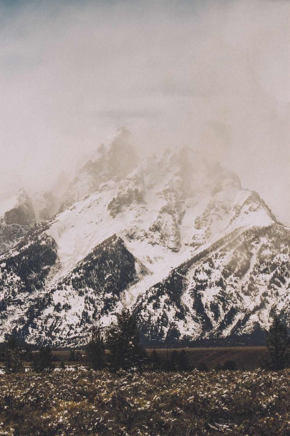 Snow-capped mountain peak shrouded in misty clouds with rugged terrain in the foreground.