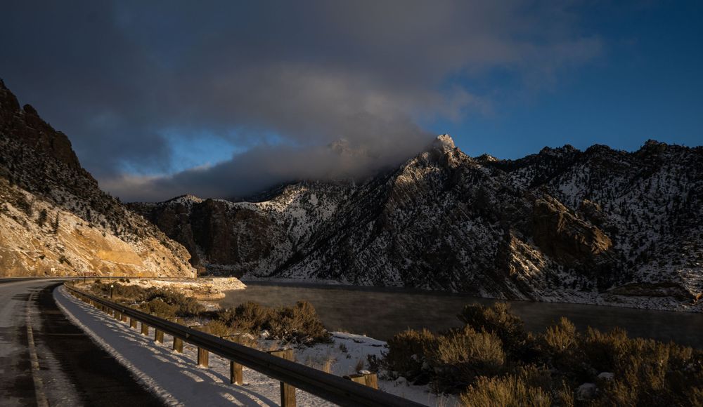 Winding mountain road covered in snow with dramatic cloudy sky and sunlight breaking through.