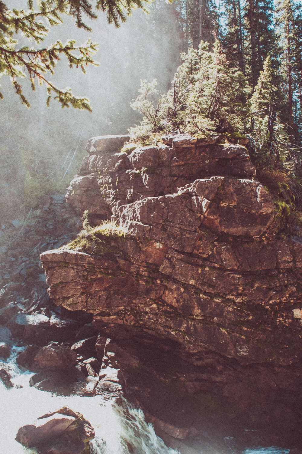 Sunlit rocky cliff face with cascading waterfall and evergreen trees in a misty forest setting.