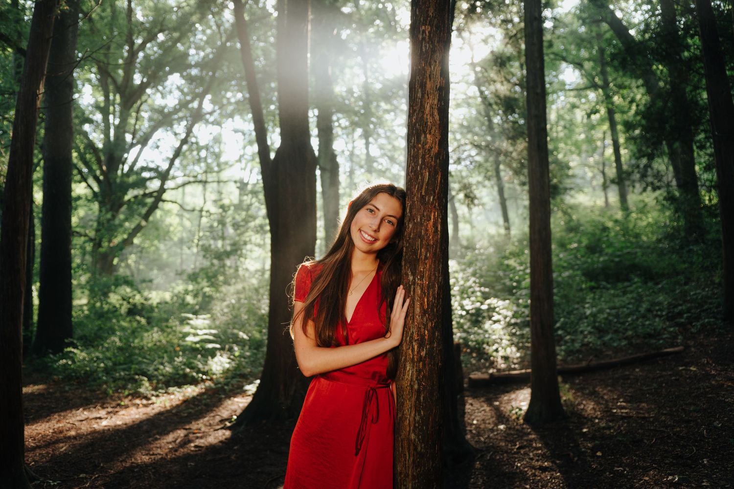 Portrait in red dress leaning against tree trunk with sunlit forest background.