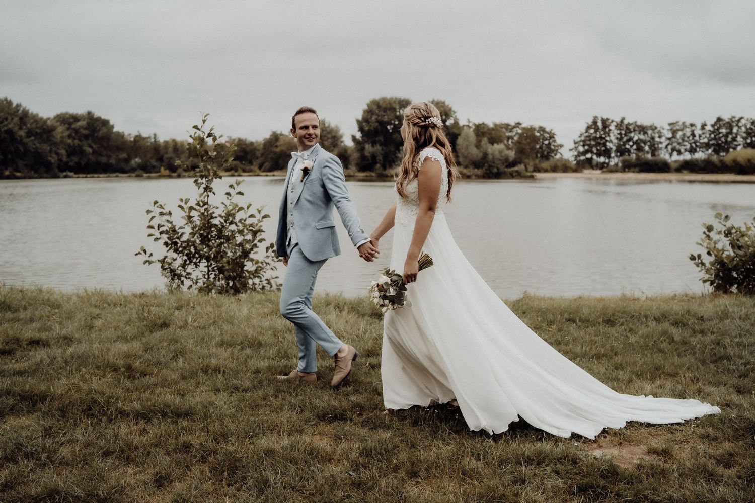 A couple walks hand in hand by a lake in wedding attire.