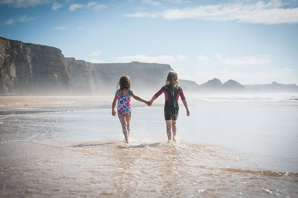 Two people holding hands while walking on a misty beach with dramatic cliffs in the background.