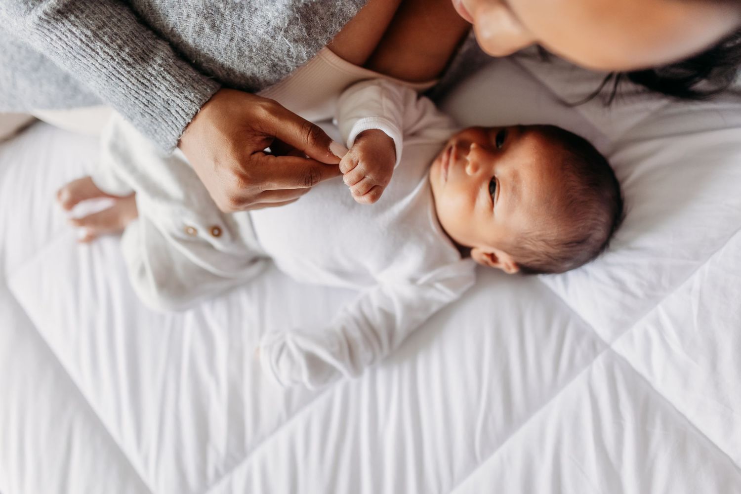 A tiny infant wearing white lies on soft bedding while a hand gently reaches to touch them.