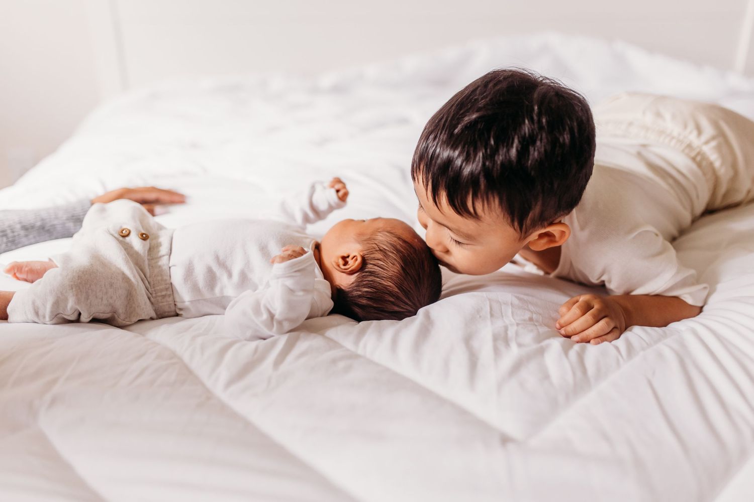 Child lying on white bed next to sleeping baby with white cat nearby.
