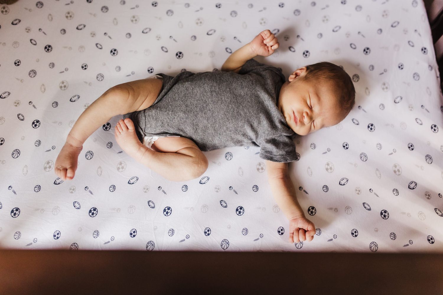 A sleeping baby in grey onesie lying on patterned bedding with arms spread out.